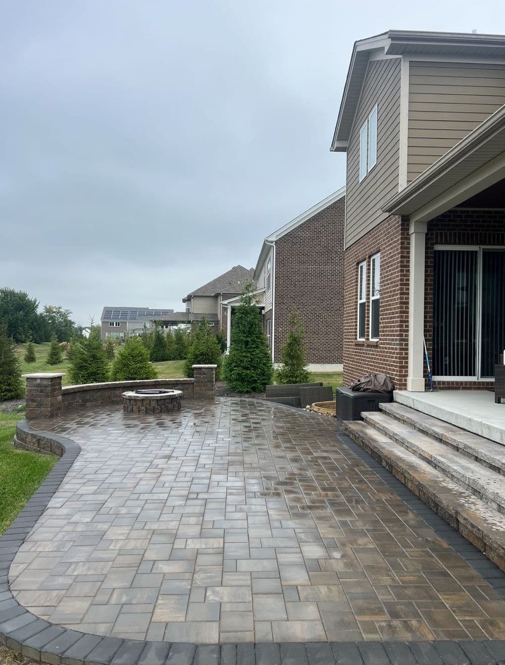 Paved backyard patio with fire pit and landscaped greenery under a cloudy sky.