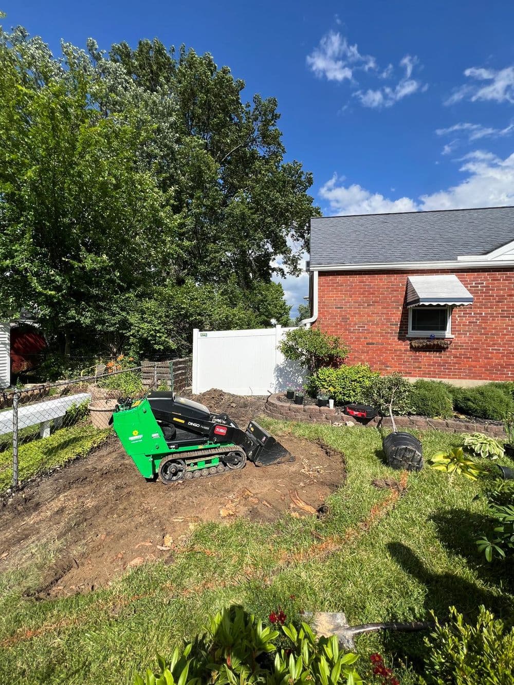 Mini excavator on lawn performing landscaping work near a brick house and fence.
