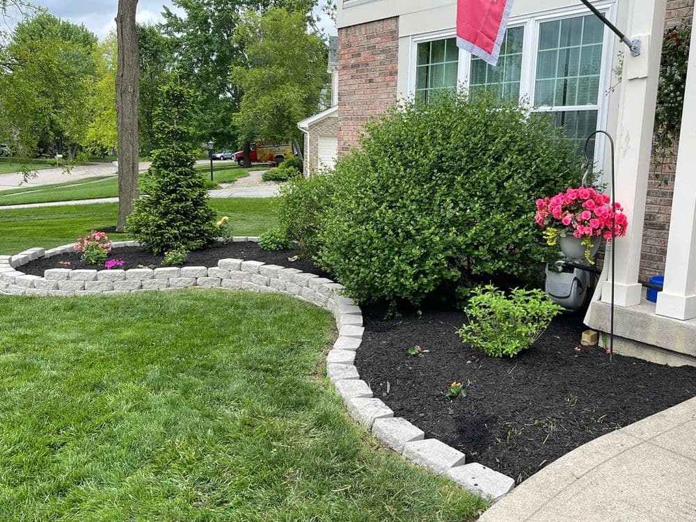 Lush garden with colorful flowers, stone border, and freshly mulched landscape near a home.