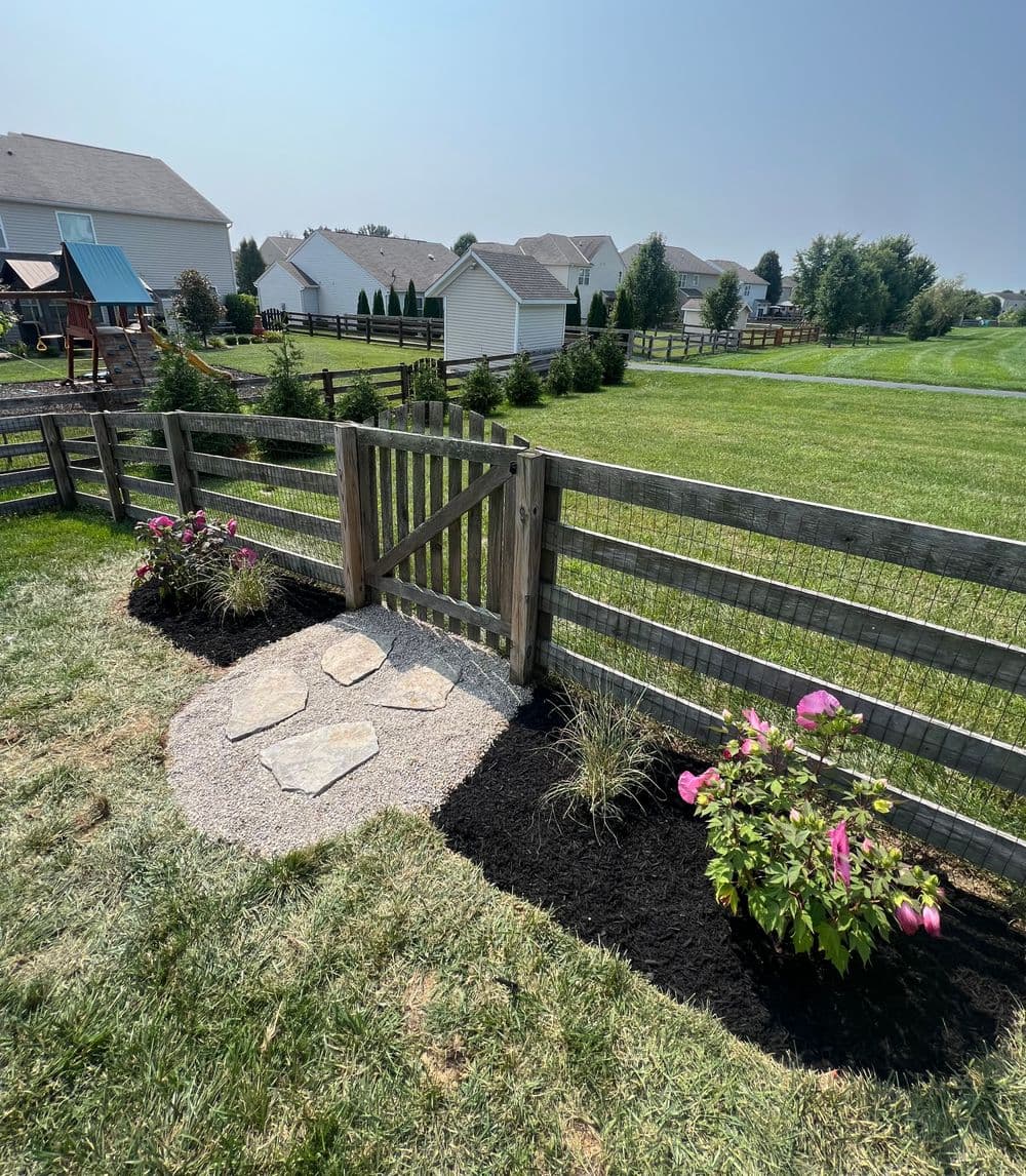 Backyard garden with stone path, wooden gate, and colorful flowers in mulch landscape.