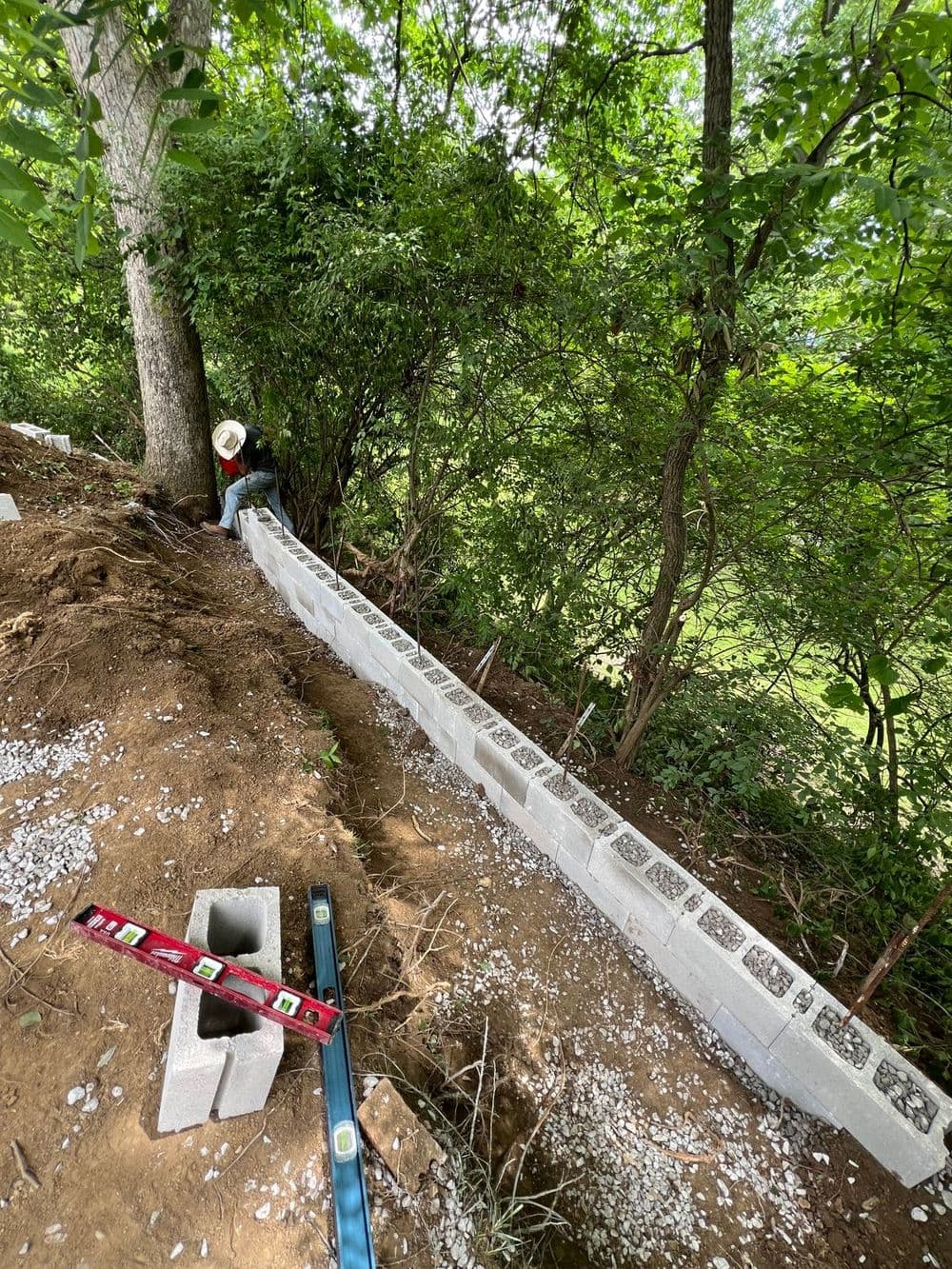 Construction worker installing a masonry wall on a slope among trees with tools nearby.