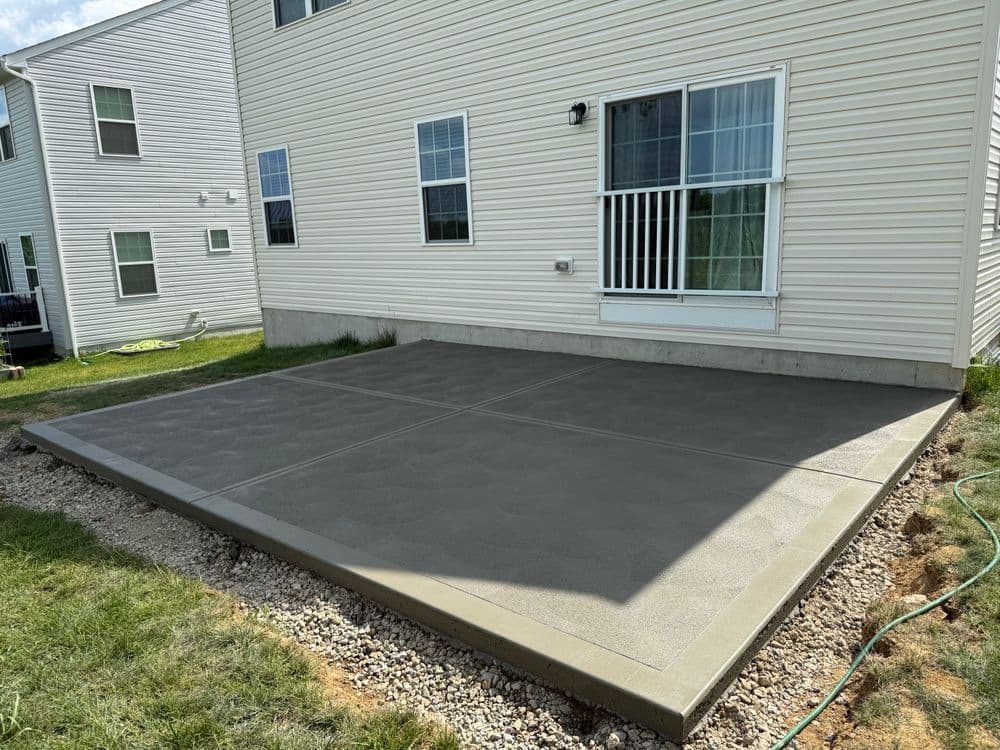 Concrete patio installation behind a house with grassy edges and a sliding door.